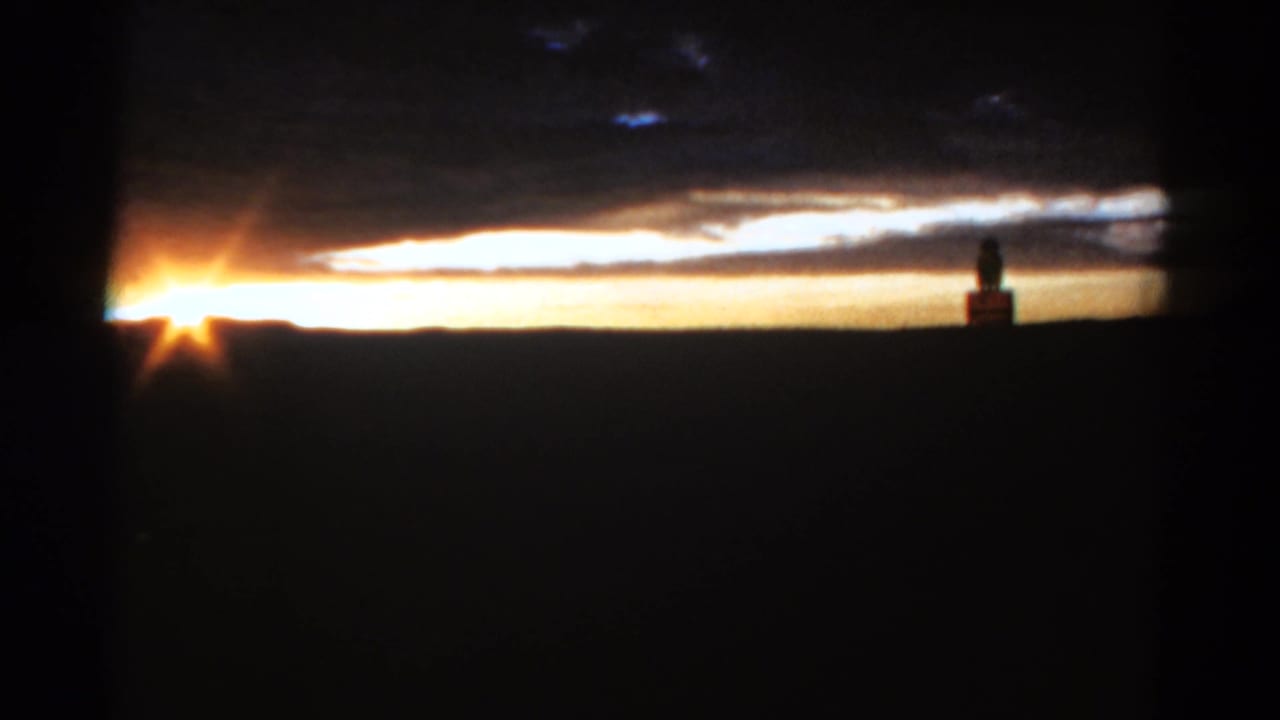 Low sun on a dark horizon with orange sky and a distant statue-like silhouette.