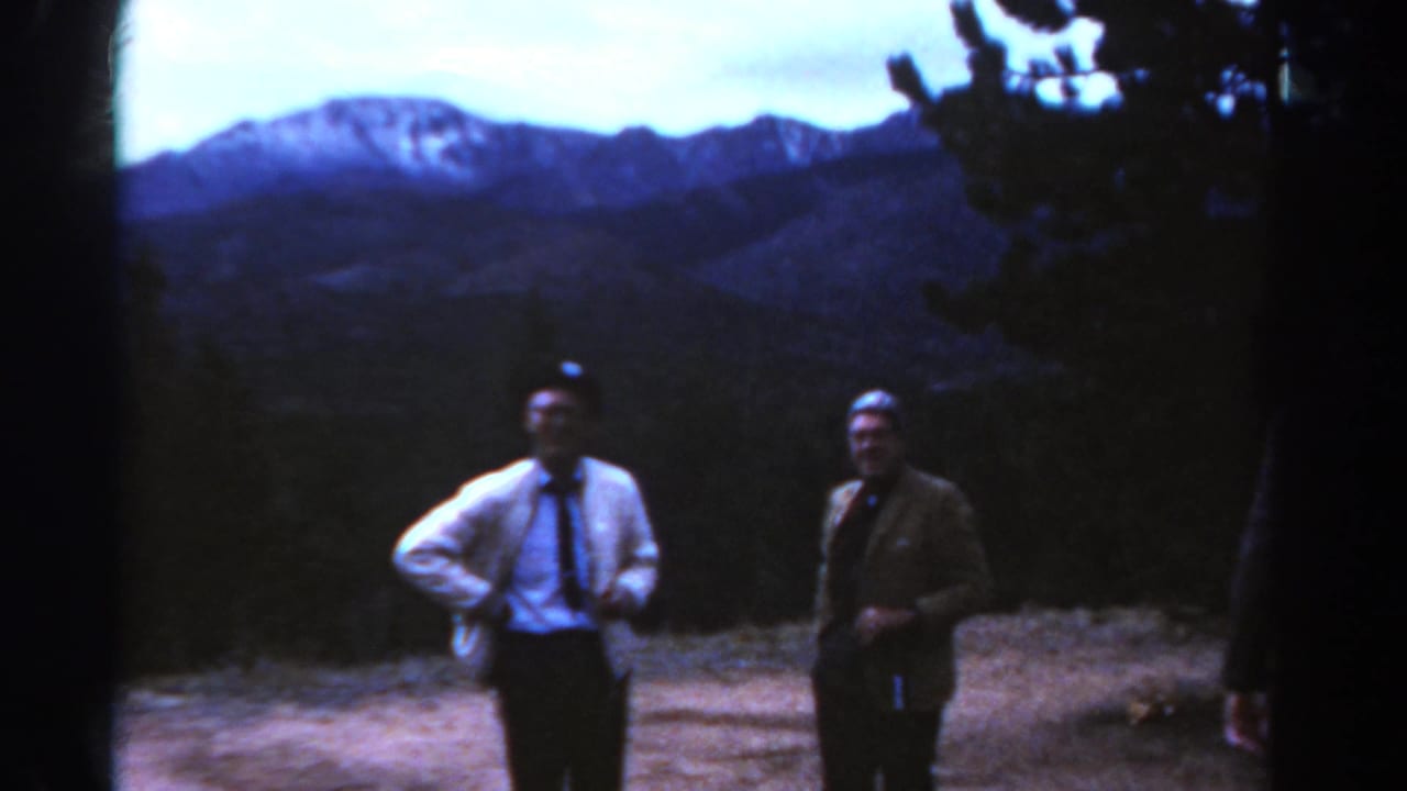 Two men standing near a mountain overlook with evergreen trees and peaks behind them.