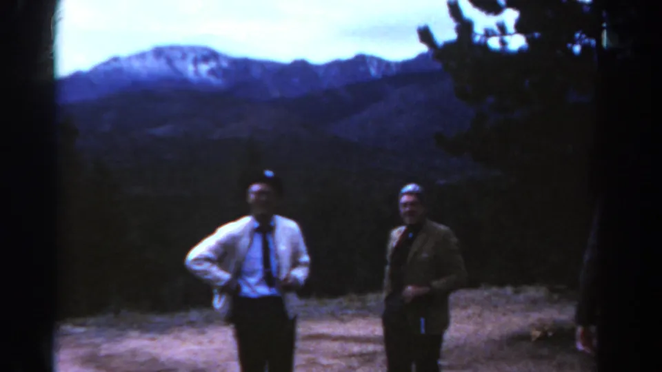 1967 Colorado 8mm frame of two men posing at a mountain overlook, with dark trees and snow-streaked peaks in the background.
