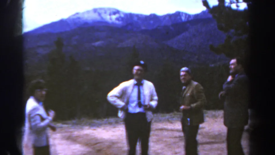 1967 Colorado home-movie frame showing three men and one woman gathered at a mountain overlook with evergreen trees and distant peaks.