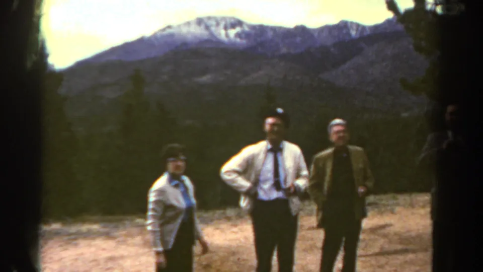 1967 Colorado 8mm frame of a woman in a light cardigan and two men in formal clothing posing before mountains and trees.