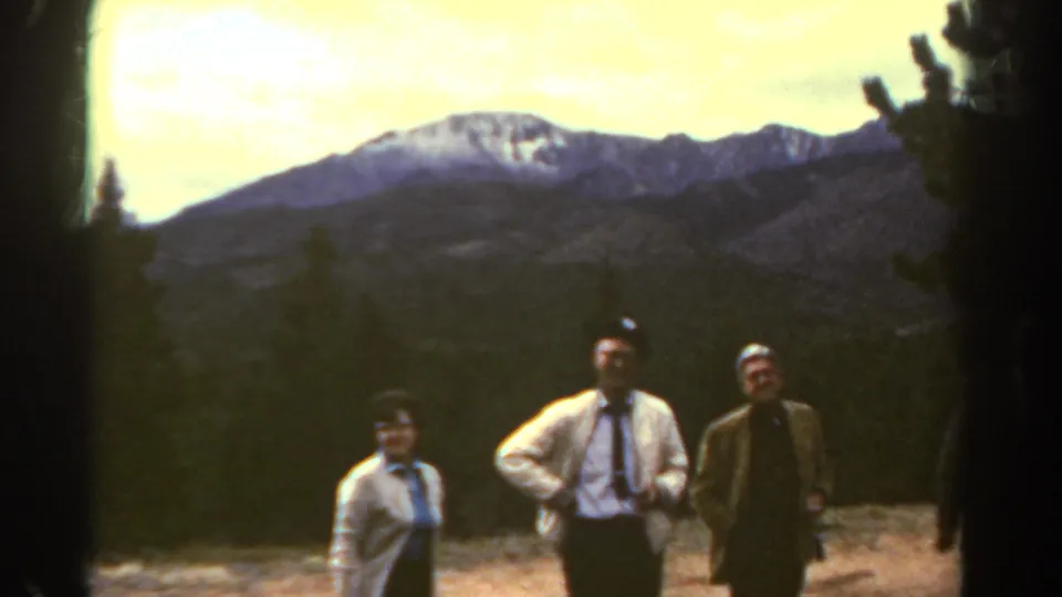 1967 Colorado home-movie frame with a woman and two men posed in front of dark trees and a broad snow-streaked mountain peak.