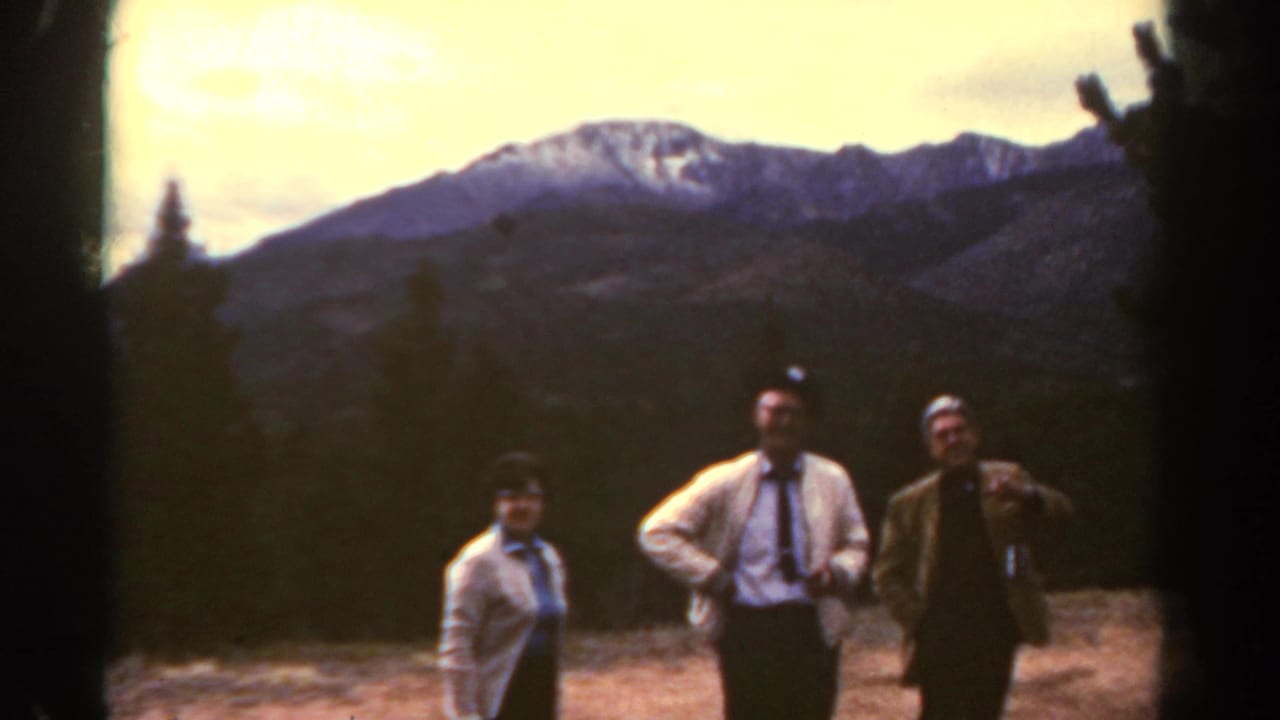 Three adults stand before evergreen trees and distant mountains.
