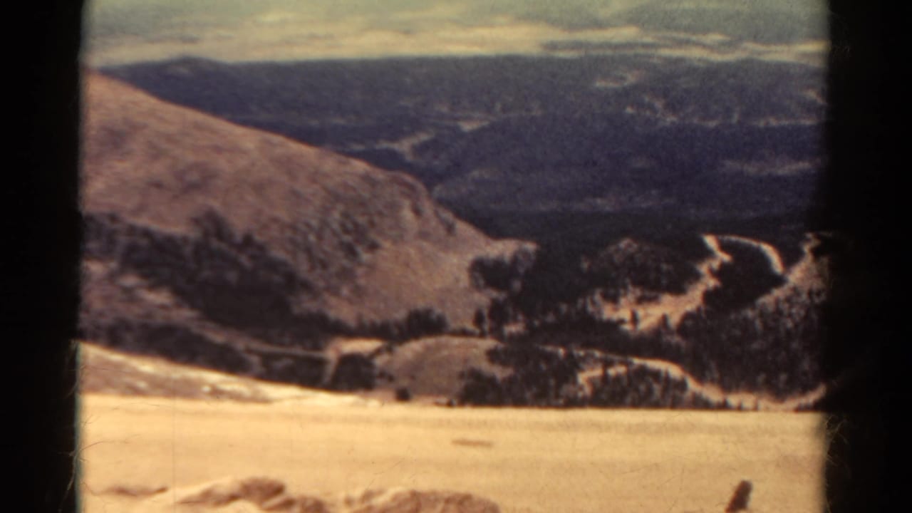 High overlook view across pale slopes, dark ridges, winding paths, and a hazy sky.