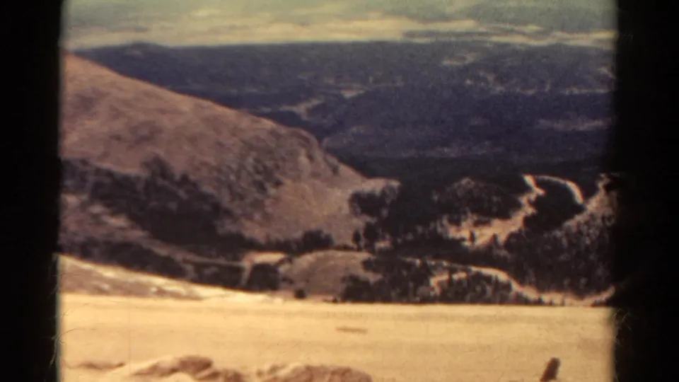 1967 Colorado 8mm frame showing a high, arid slope above dark mountain ridges and winding roads under a hazy sky.