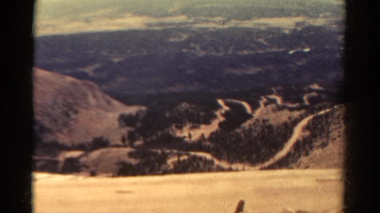 Panoramic mountain view with pale hillside, dark valley ridges, and winding road traces.