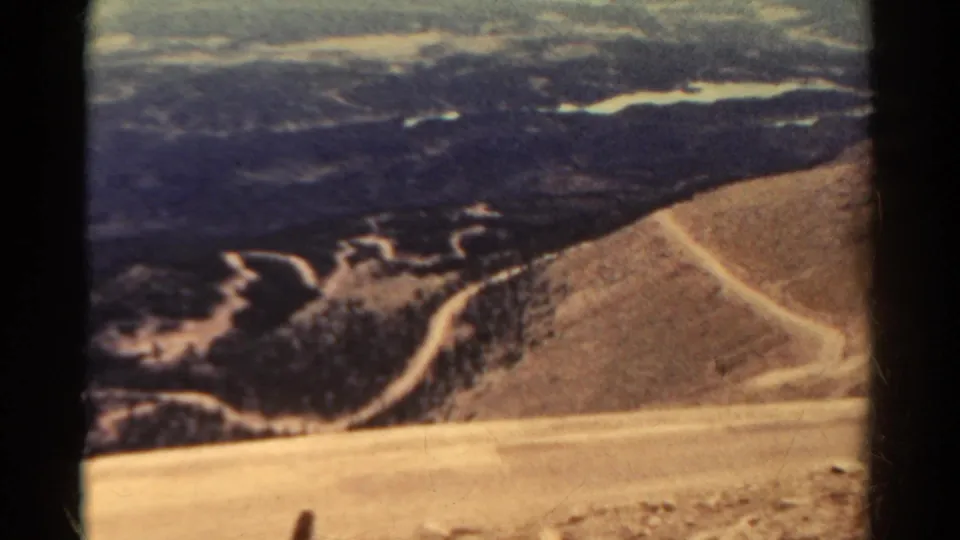 1967 Colorado 8mm frame showing a pale open slope, a curving road, dark mountain ridges, and hazy distant terrain.