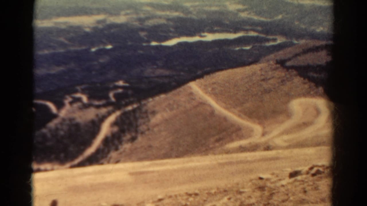 Curving dirt road crosses a dry hillside with dark scrub and distant terrain beyond.