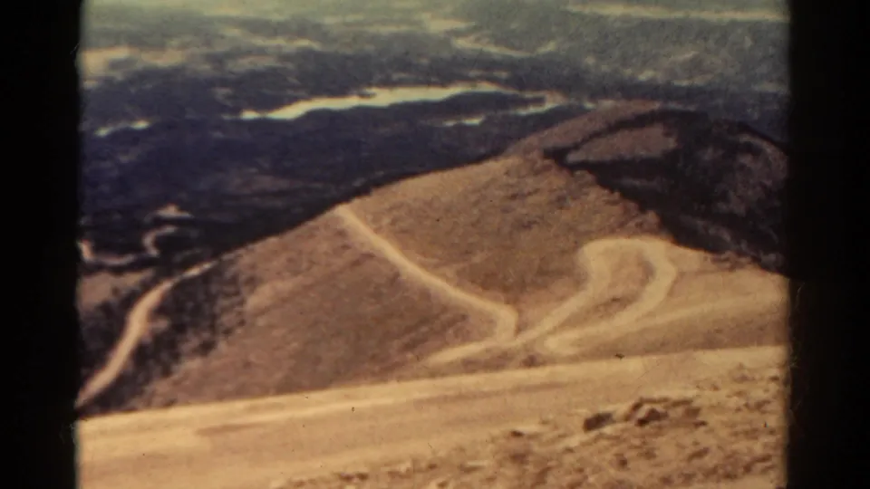 Vintage 8mm Colorado frame showing a pale dirt switchback on a sandy slope with darker vegetation and distant hills.