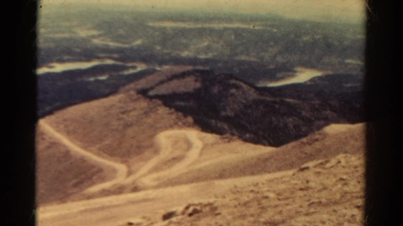 Barren mountain slope with winding dirt trails and distant hazy ranges beyond.