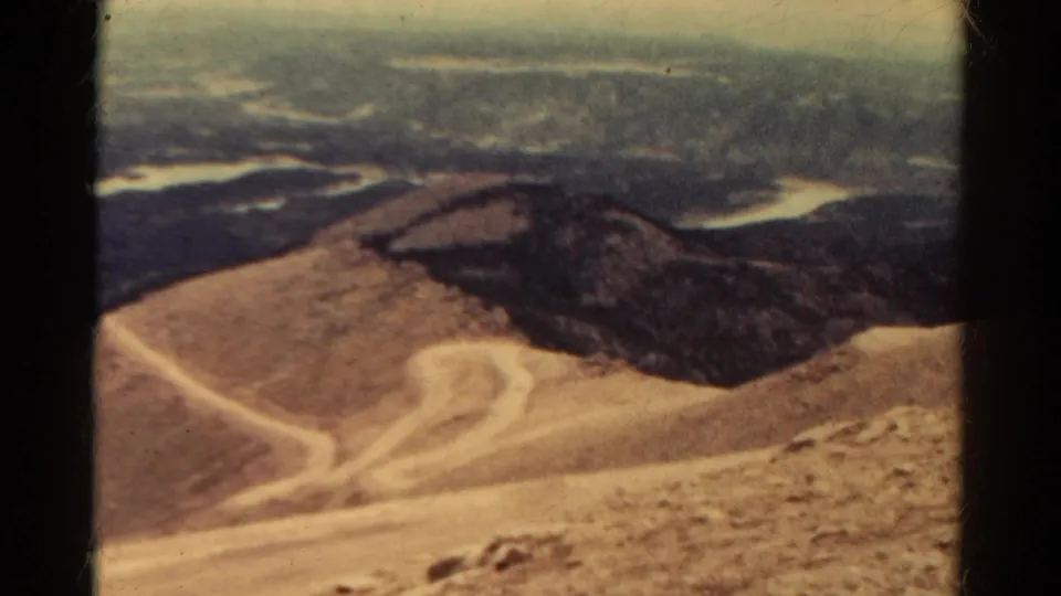 1967 8mm Colorado home-movie frame showing arid mountain slopes, pale trails, and distant ranges under a cloudy sky.