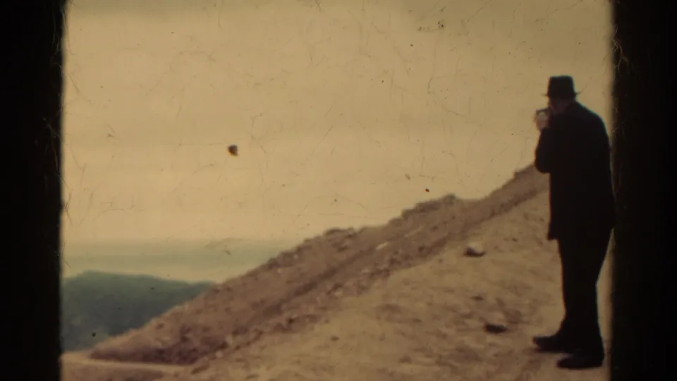 1967 Colorado 8mm frame showing a man in a dark suit and hat filming from a dusty mountain overlook.