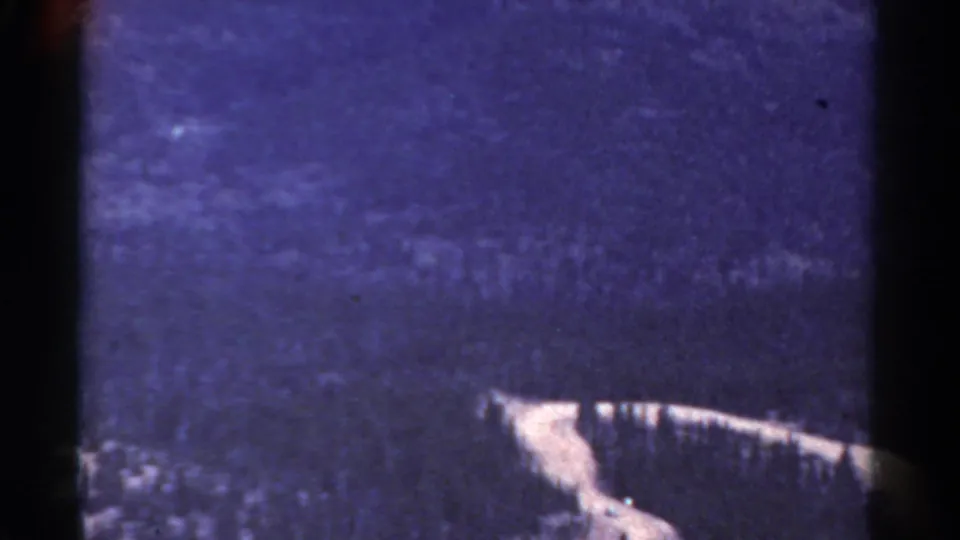 1967 Colorado home-movie still showing a blue-purple mountainside, soft distant forest texture, and a pale diagonal ridge or road line below.