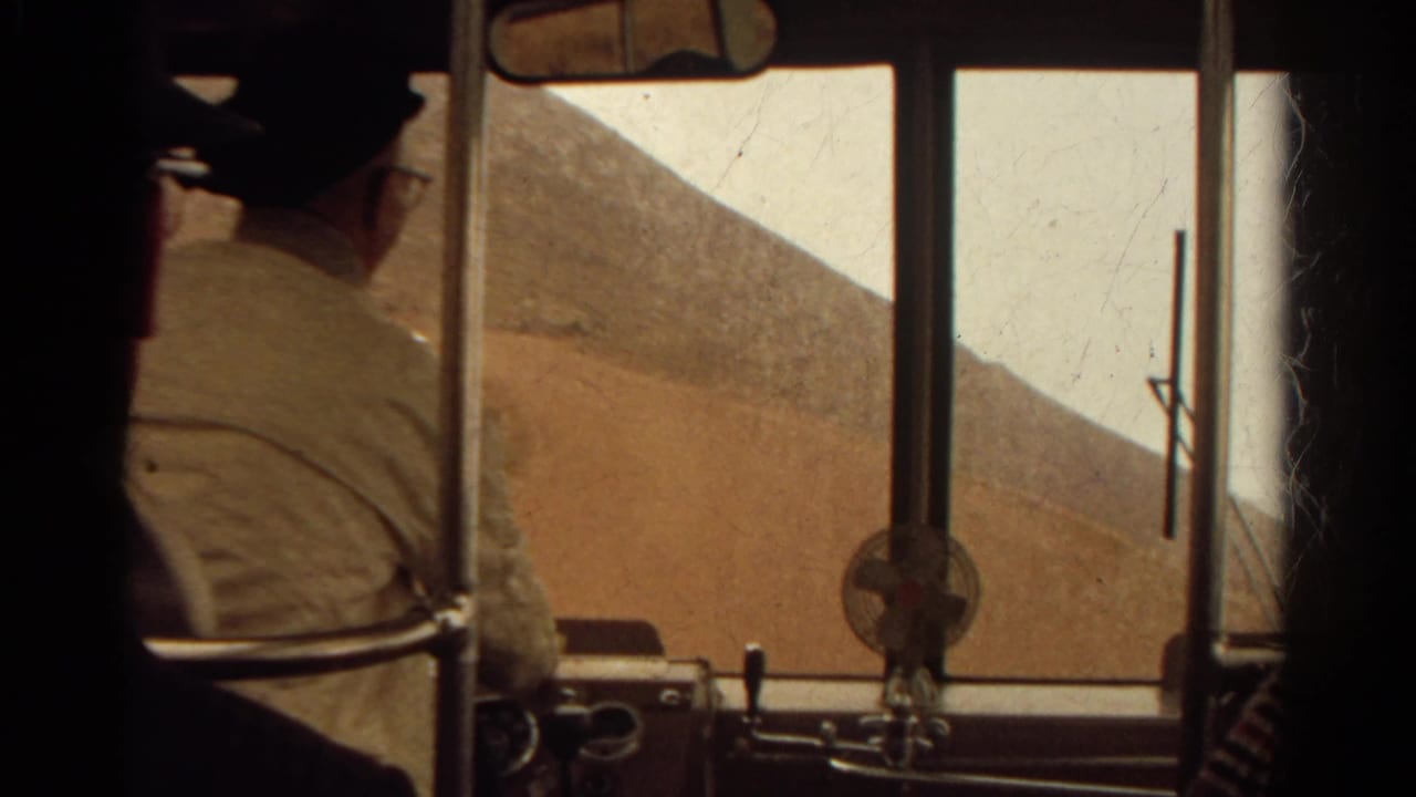 Inside a 1967 Colorado bus, an adult passenger in a light shirt sits near the front as a dry hillside passes outside.