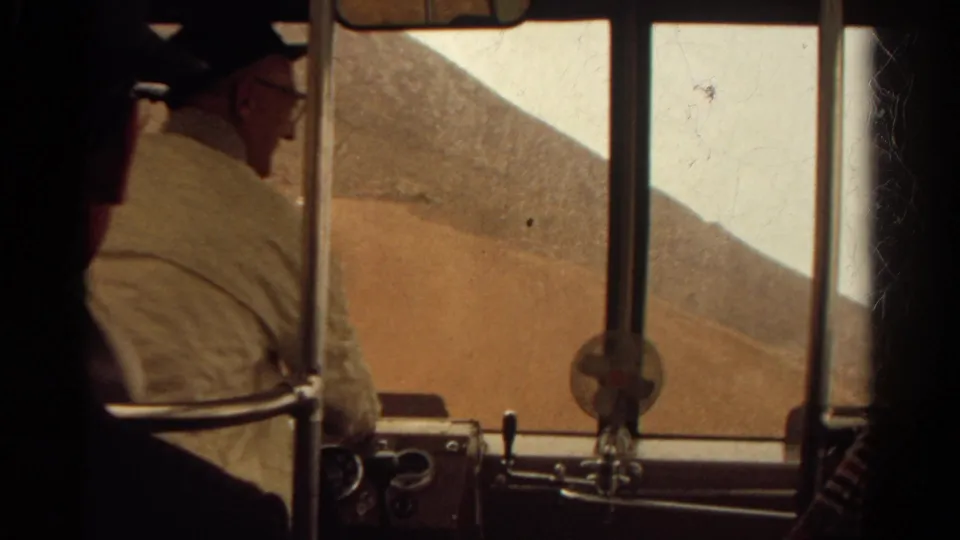 A home-movie view from inside a Colorado bus shows a seated passenger, metal rails, dashboard details, and the arid hillside beyond the window.