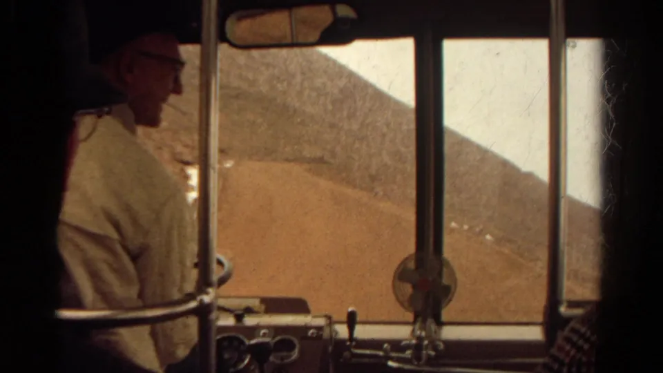 The final frame holds on the bus front interior, with an adult passenger in profile, dashboard fan, window rails, and dry Colorado hillside outside.