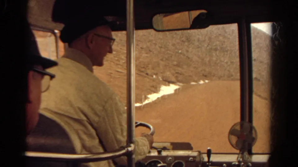 Passenger-side view of the driver, windshield posts, and dashboard as the vehicle travels a Colorado dirt road in 1967.