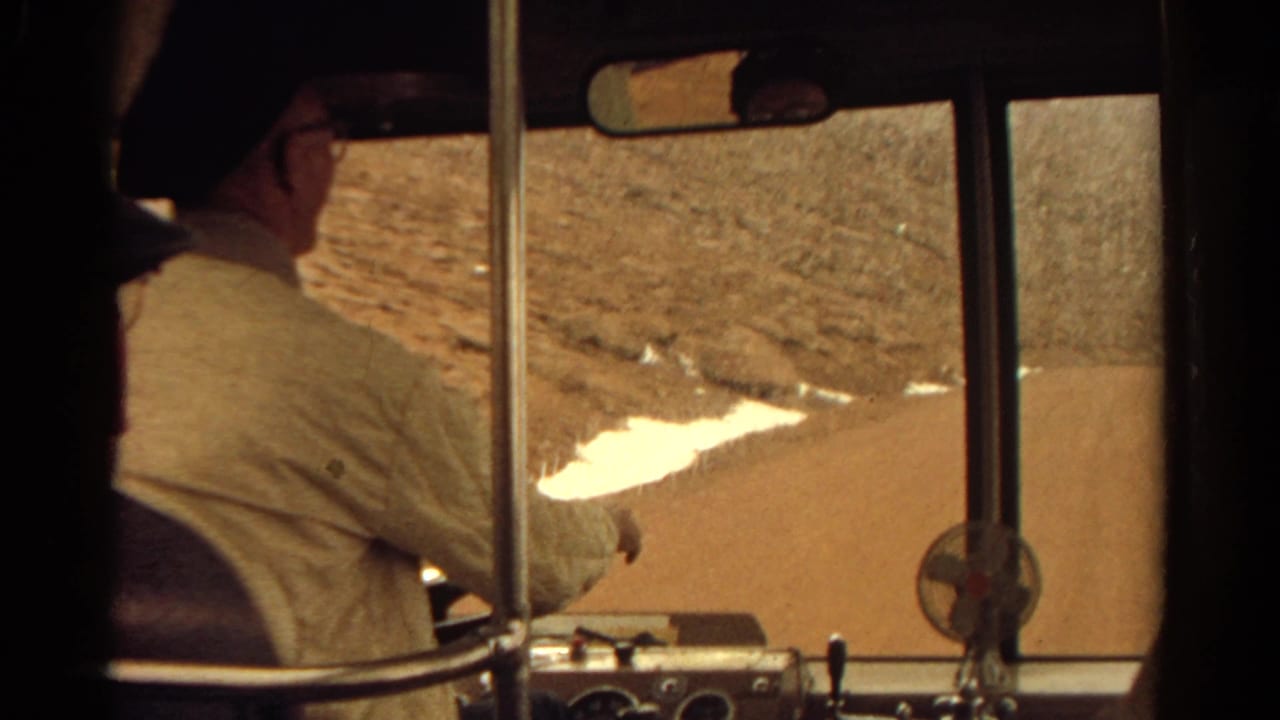 Bus driver in profile reaches forward as the windshield frames a road beside rocky terrain.