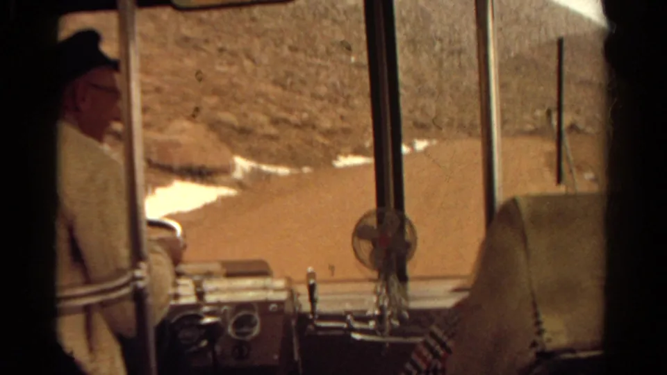 Final 1967 Colorado bus interior frame showing the driver at left, dashboard instruments, windshield supports, and arid terrain with snow traces.