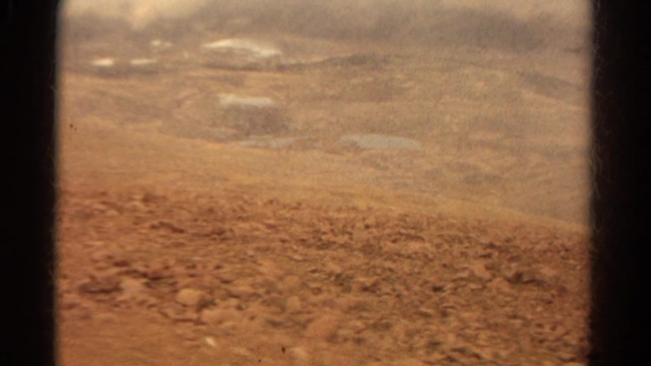 1967 Colorado home-movie frame looking from inside a vehicle across reddish dirt, scattered rocks, and hazy distant ground.