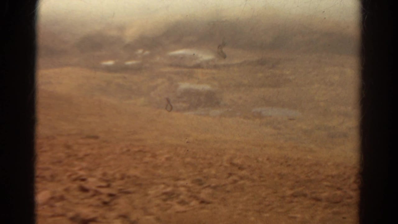 Faded 8mm view from inside a vehicle across dry Colorado terrain with rocks, haze, and faint distant shapes.