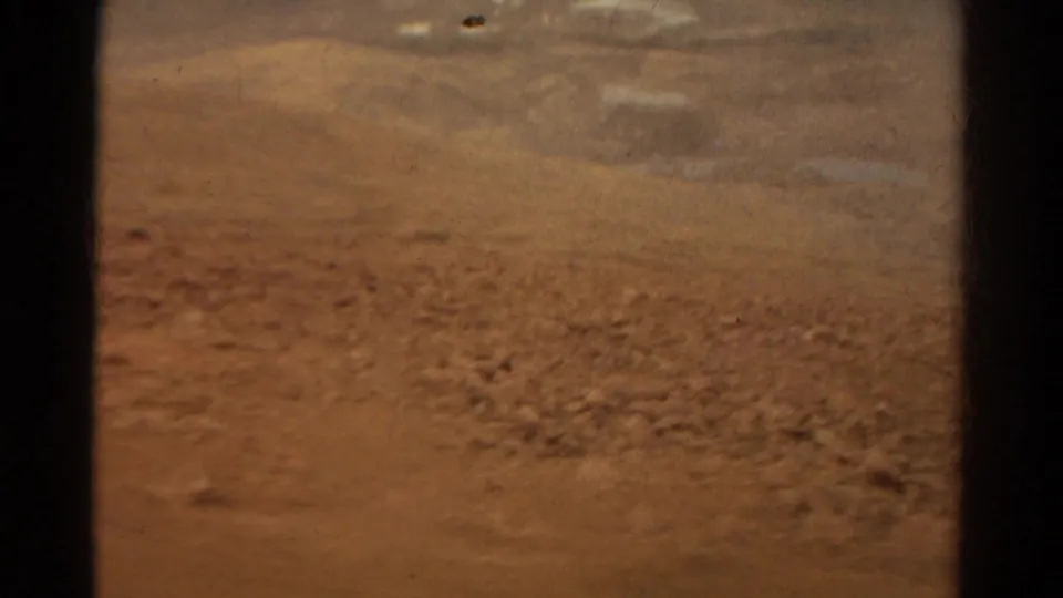 Soft 8mm frame from a moving vehicle showing rough reddish soil, scattered stones, and a blurred dry background in Colorado.