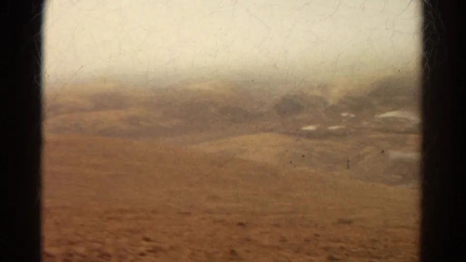 Faded Colorado road-view frame with dry open ground, a dark window divider, haze, and faint distant structures or mounds.
