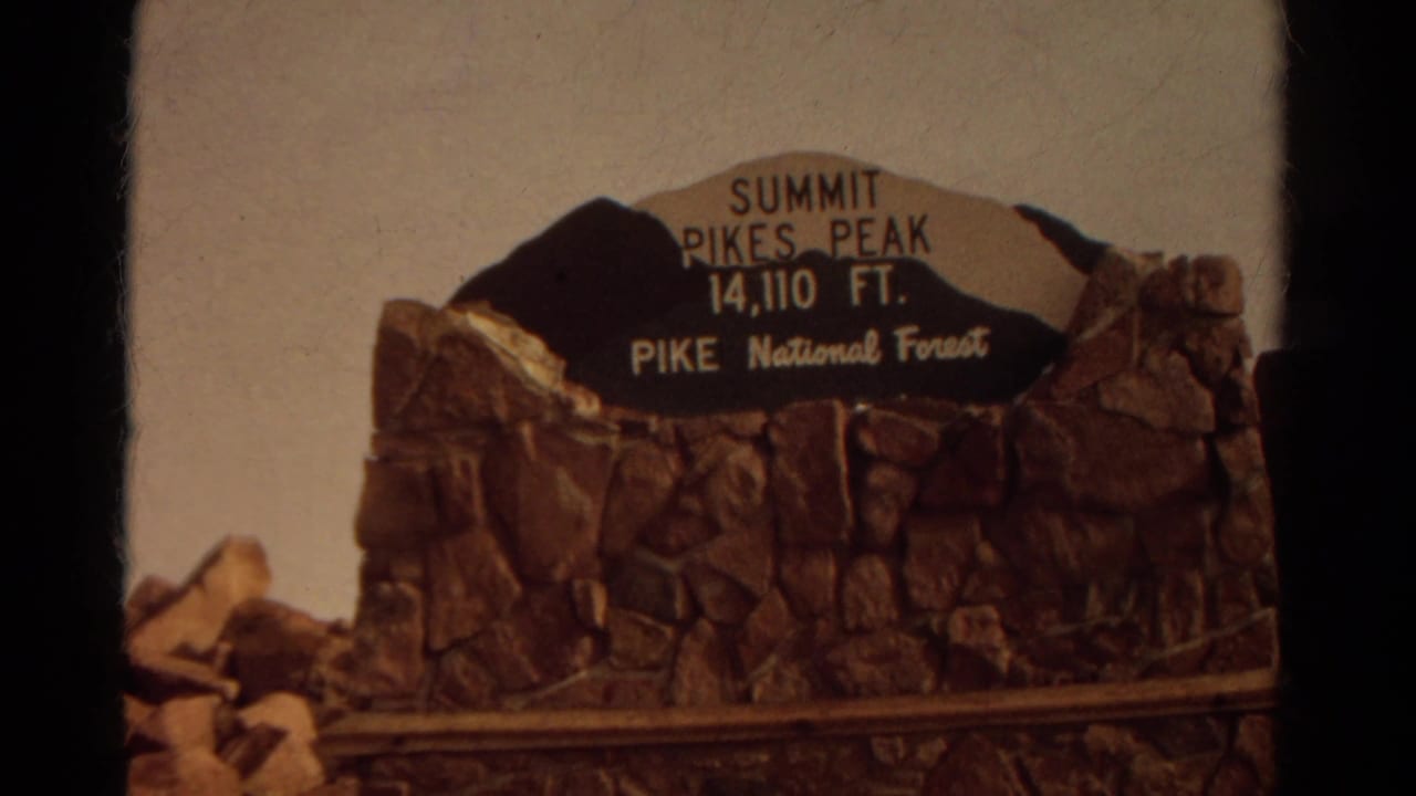 Pikes Peak summit sign reading 14,110 feet in Pike National Forest, framed on 1967 home-movie footage.