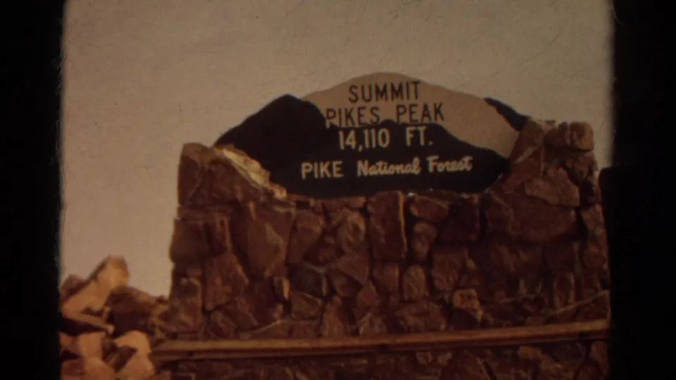 Pikes Peak summit sign reading 14,110 feet in Pike National Forest, framed on 1967 home-movie footage.