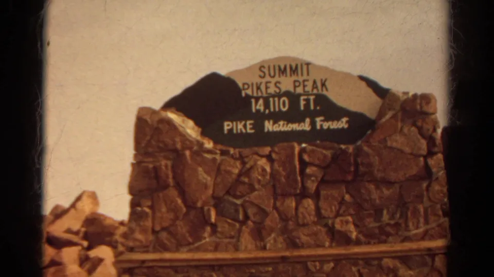 Centered view of the Pikes Peak summit sign and stone wall, with a partial seated visitor at the lower left edge.