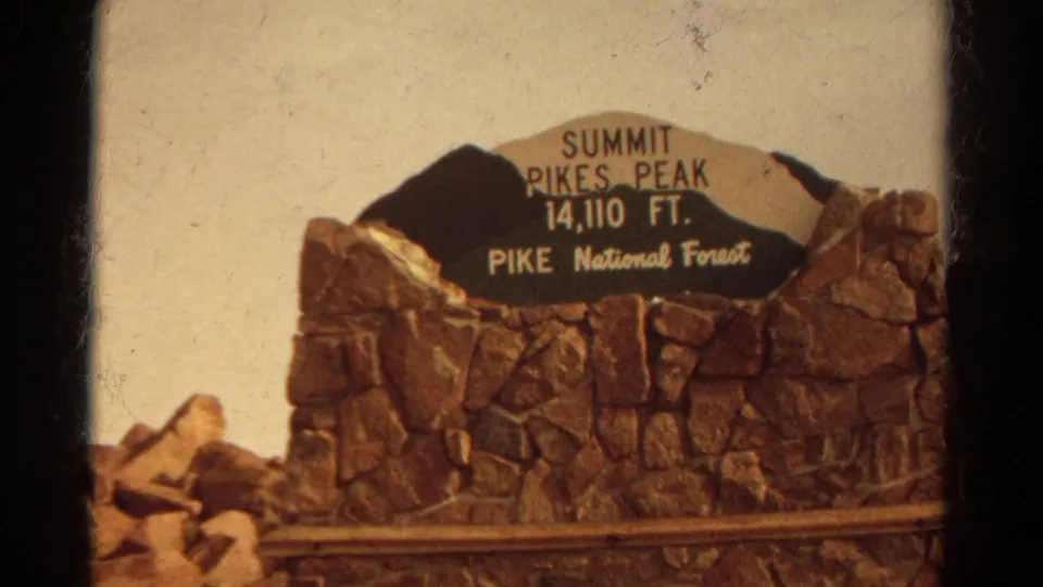 Warm 8mm view of the Pikes Peak summit sign, stone base, rail, rocks, and pale sky in Colorado.