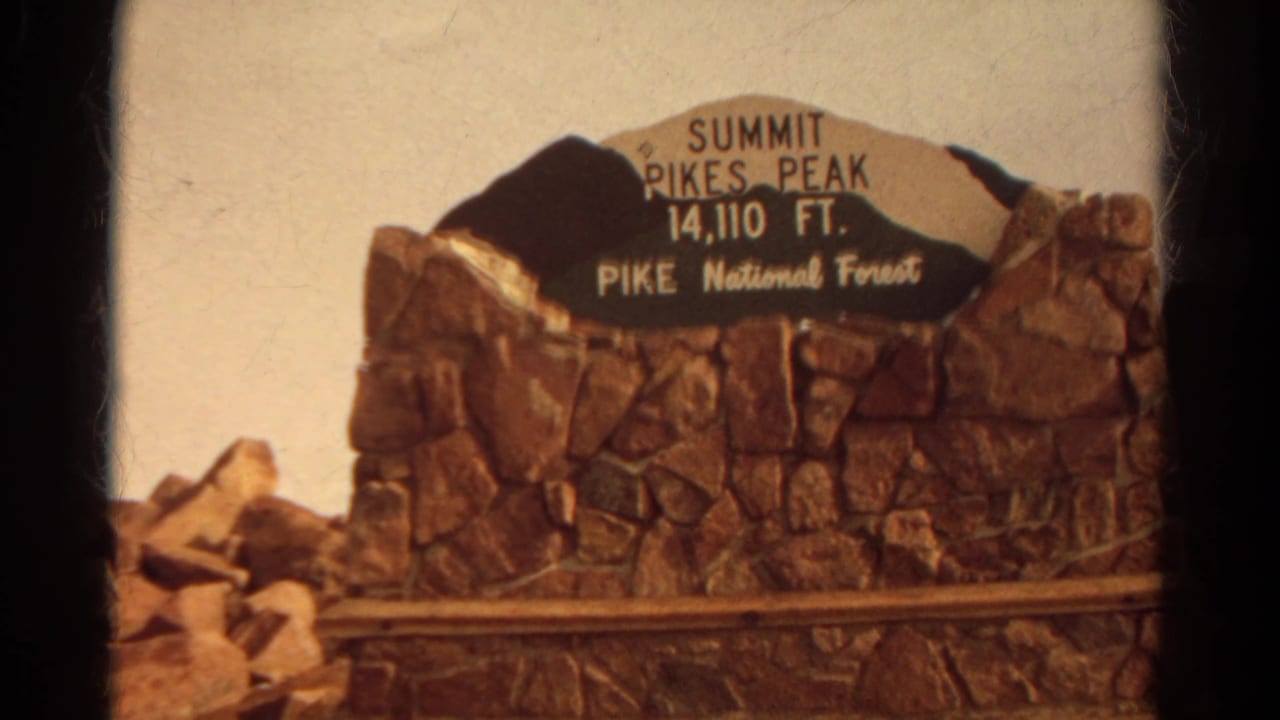 Close 8mm view of the Pikes Peak summit marker in Pike National Forest, with white lettering on a dark sign panel.