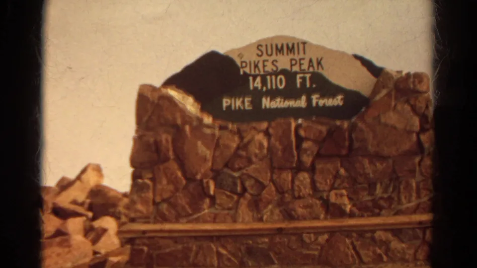 Close 8mm view of the Pikes Peak summit marker in Pike National Forest, with white lettering on a dark sign panel.