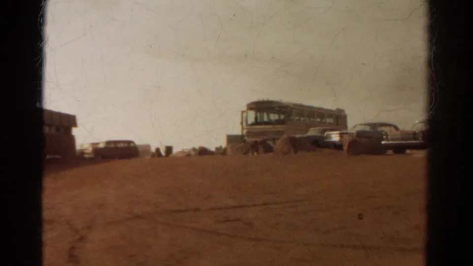 8mm frame of the Pikes Peak summit parking area, showing a bus and cars spaced across a dusty foreground.