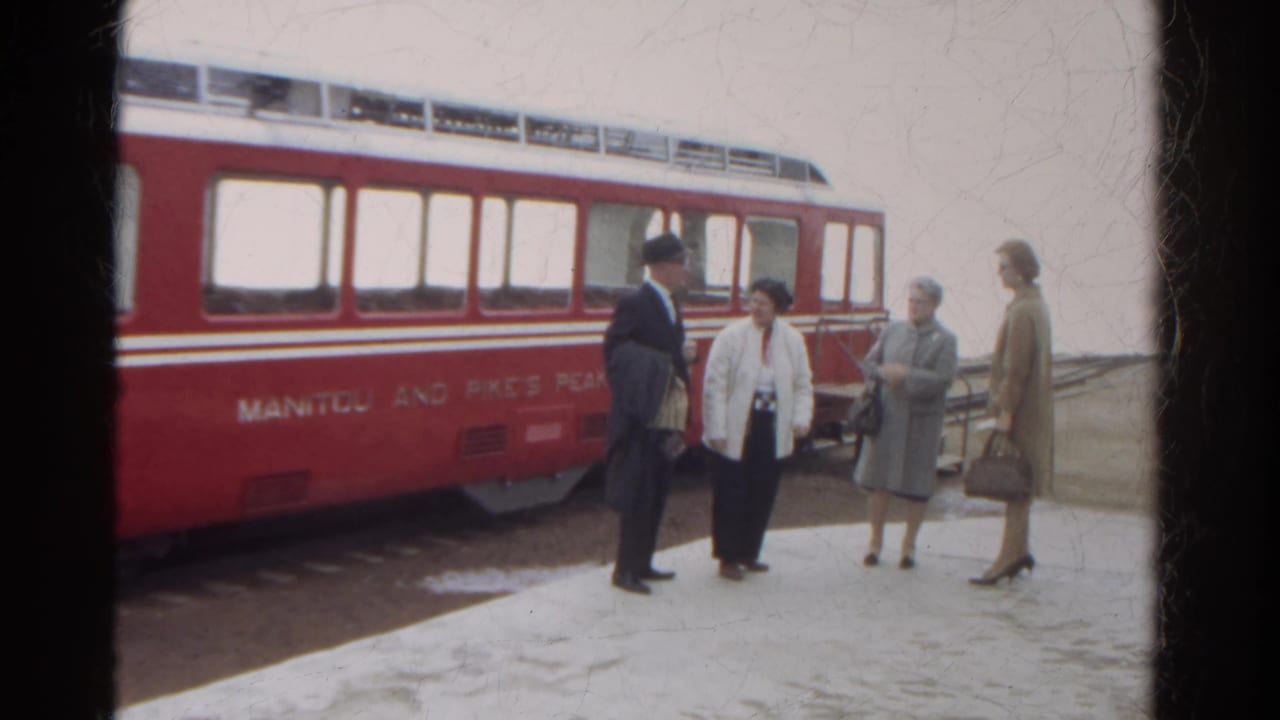 In 1967 Colorado, three women and a suited man stand talking near a red Manitou and Pike's Peak railway car on a snowy platform.