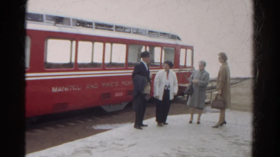 In 1967 Colorado, three women and a suited man stand talking near a red Manitou and Pike's Peak railway car on a snowy platform.