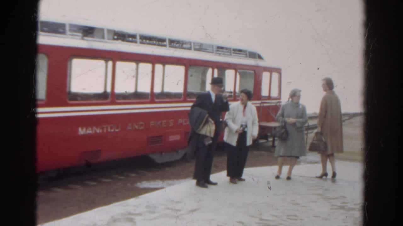 A suited man gestures while three women stand beside the red railway car.