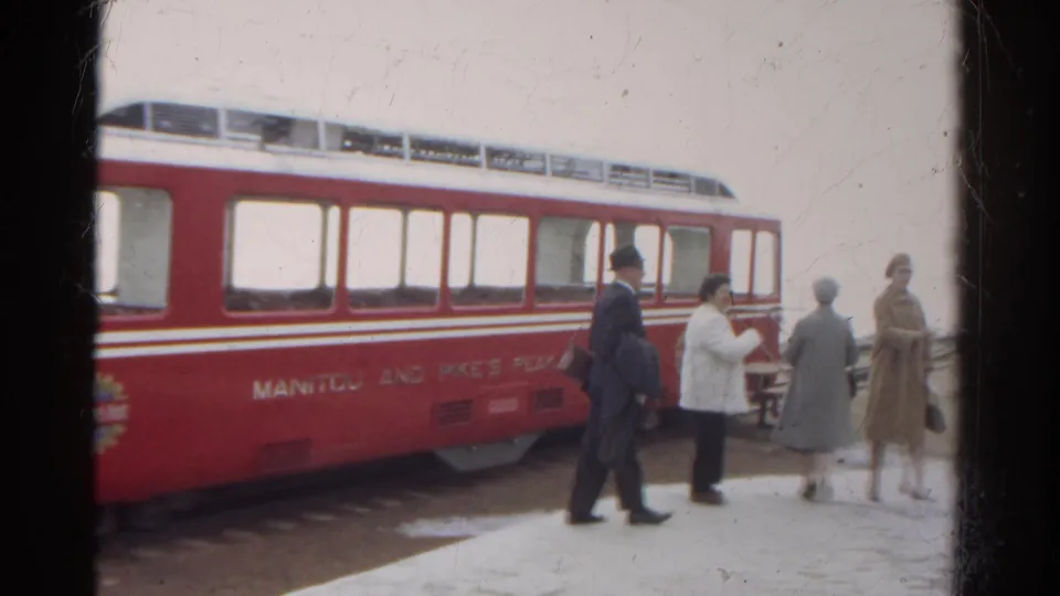 The suited man walks left while the women move along the snowy platform beside the red Manitou and Pike's Peak railway car in Colorado.