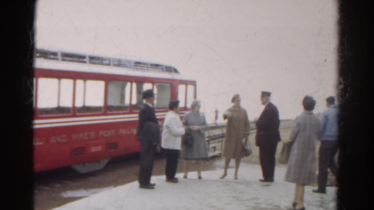 Adults in 1960s winter clothing stand and converse near a red Manitou and Pike's Peak Railway car in Colorado.