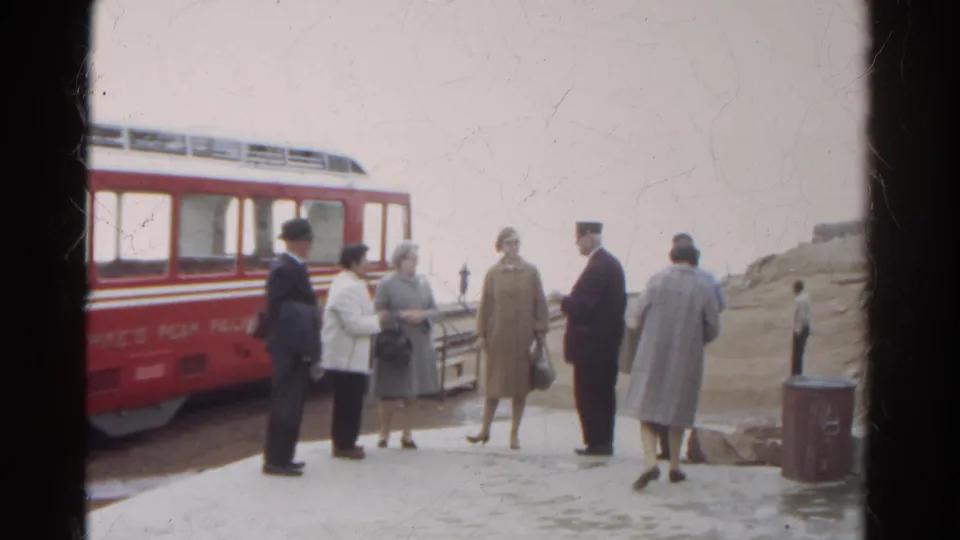 Colorado mountain railway platform scene with adults, a red rail car, winter coats, and pale terrain in the background.
