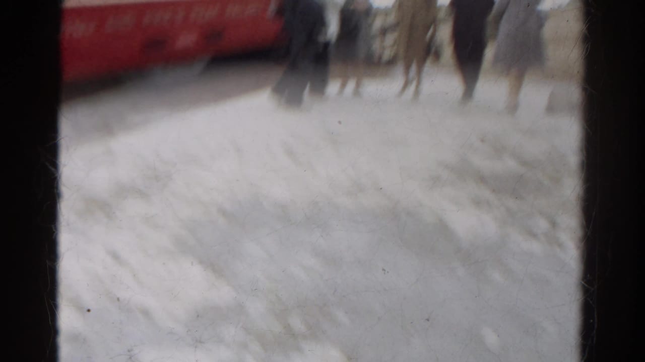Motion-blurred 1967 Colorado home-movie frame with a red railcar at left and pedestrians near a light platform surface.