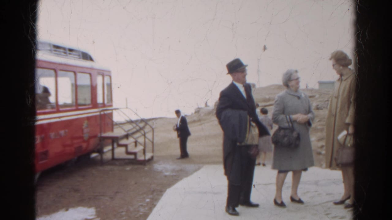 Adults in coats stand near a red railway car and boarding steps.