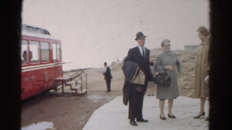 1967 Colorado frame showing a suited man and women in coats beside the red mountain railway car and snowy platform edges.