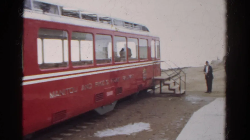 Red Manitou and Pike's Peak Railway car with white trim, boarding steps, and a lone man on the platform in Colorado.