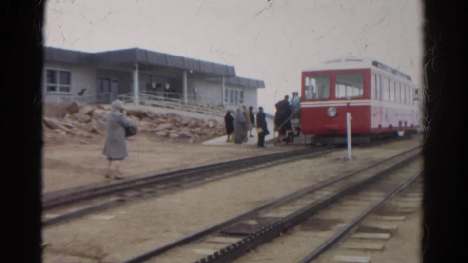 Passengers in coats gather by the tracks as a red mountain railway car waits near a station building in Colorado archival footage.