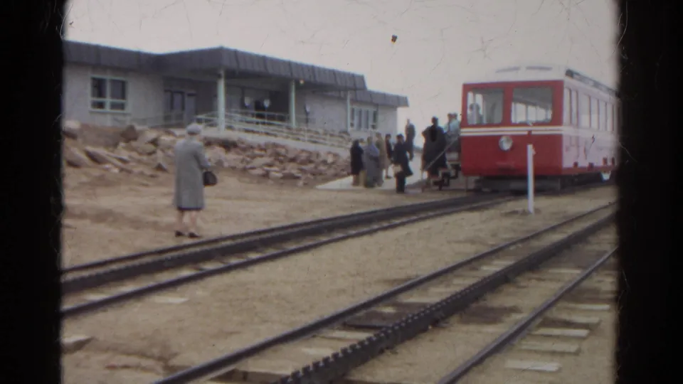 A red Manitou and Pike's Peak Railway car waits as passengers board beside multiple tracks at a mountain station.