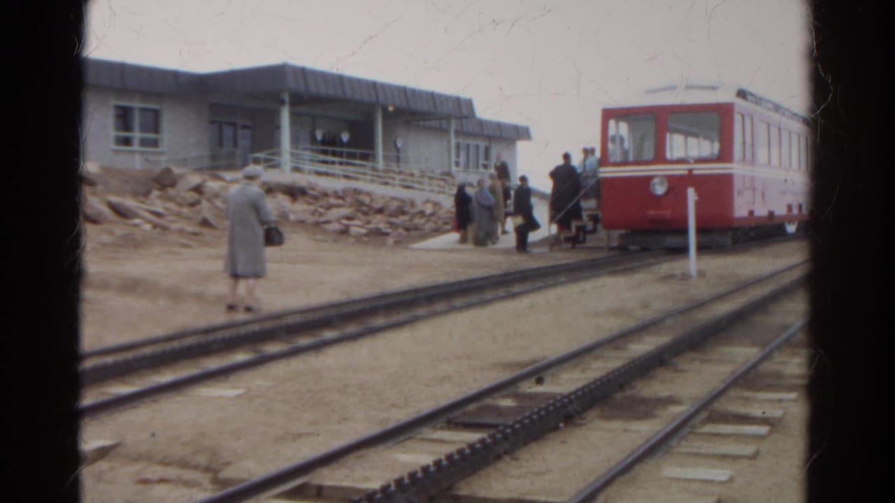 Red railcar at a small station with people near tracks on an overcast day.