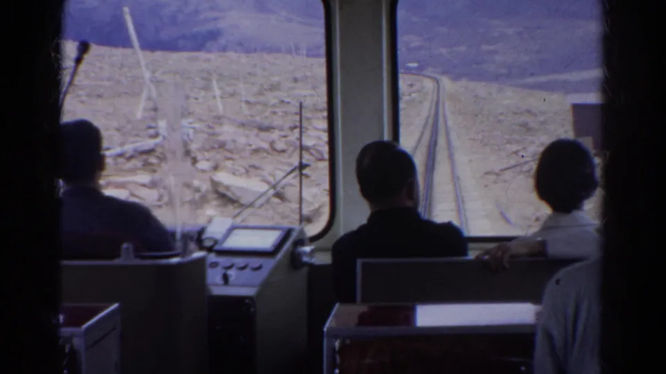 Interior 1967 Colorado travel view with seated passengers facing a front window and narrow tracks through dry landscape.