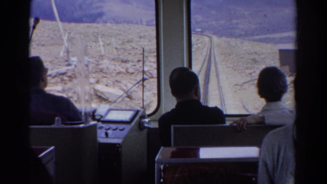 Passengers sit inside a train looking through large windows at rocky terrain and tracks ahead.