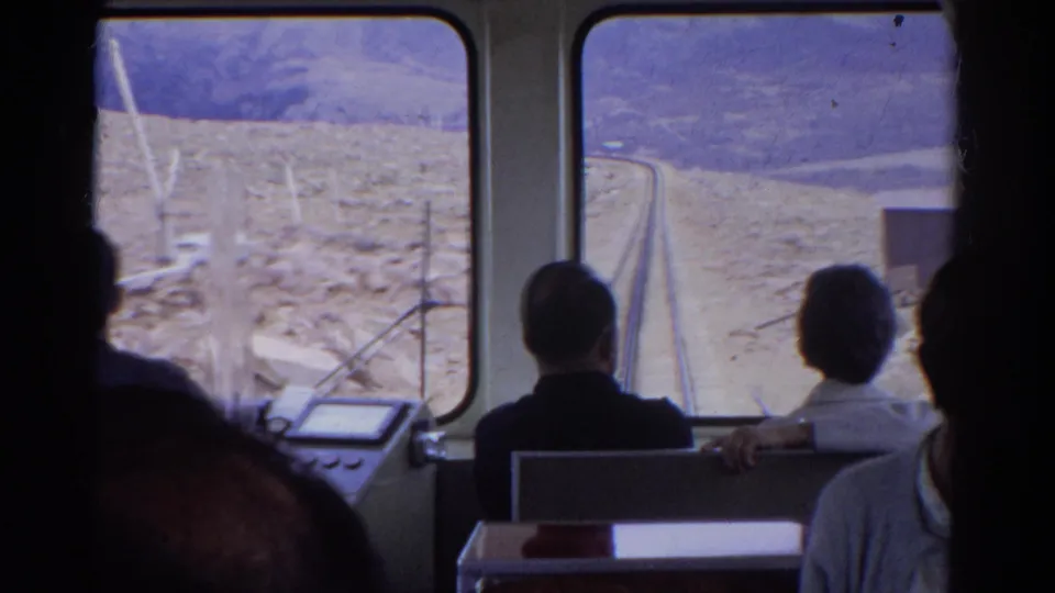 Interior passenger view from a 1967 Colorado train, with travelers facing the windows and mountain tracks ahead.
